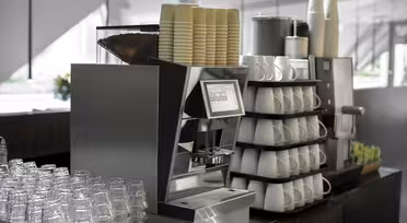 Et moderne kaffemaskinoppsett med stabler av papirkopper på toppen. Glass og hvite keramiske kopper er pent arrangert i hyller. En organisert visning av tepakker er sett i nærheten, mot et bakteppe av et lyst, luftig rom.