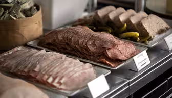 En delikatessedisk som viser brett med oppskåret delikatessekjøtt inkludert skinke og salami, ledsaget av sylteagurk og paté. En liten kurv fylt med innpakkede porsjoner er synlig i bakgrunnen.