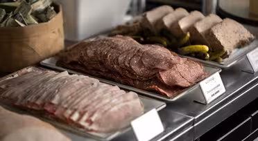 En delikatessedisk som viser brett med oppskåret delikatessekjøtt inkludert skinke og salami, ledsaget av sylteagurk og paté. En liten kurv fylt med innpakkede porsjoner er synlig i bakgrunnen.