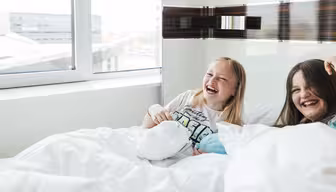 To jenter ler og sitter på en seng dekket med hvite tepper. De er i et lyst rom med et stort vindu, og en jente holder et kosedyr. Scenen er letthjertet og munter.