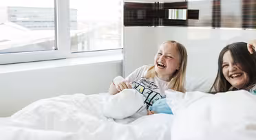 To jenter ler og sitter på en seng dekket med hvite tepper. De er i et lyst rom med et stort vindu, og en jente holder et kosedyr. Scenen er letthjertet og munter.