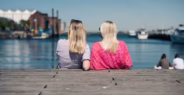 To personer med langt hår sitter på en trebrygge med utsikt over en rolig vannmasse med båter og industribygg i bakgrunnen. Personen til venstre har en lilla topp, og den til høyre har en rosa topp. Det er en solrik dag.