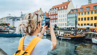En person med gul ryggsekk tar et bilde av fargerike bygninger langs en kanal i København. Båter ligger til kai i nærheten, og folk er samlet på utekafeer under overskyet himmel.
