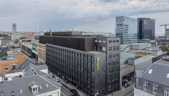 Luftfoto av et bybilde med en moderne, mørkfarget hotellbygning med navnet «Wakeup» på siden. Bebyggelsen rundt inkluderer eldre bygninger med grå tak, moderne glassbygninger og delvis skyet himmel.