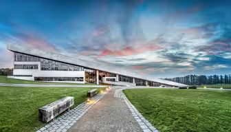 Moderne arkitektonisk bygg med unikt skråtak og store vinduer. Den ligger i et gresskledd landskap med en asfaltert sti som fører til inngangen. Himmelen er dramatisk med rosa og blå nyanser ved solnedgang.