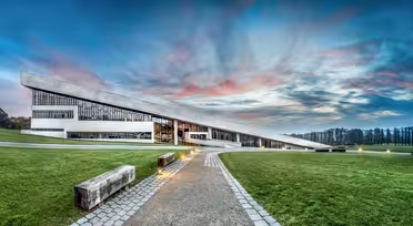 Moderne arkitektonisk bygg med unikt skråtak og store vinduer. Den ligger i et gresskledd landskap med en asfaltert sti som fører til inngangen. Himmelen er dramatisk med rosa og blå nyanser ved solnedgang.