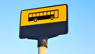 Et bussholdeplassskilt med gul bakgrunn og svart bussikon på toppen. Den nedre delen av skiltet er svart. Skiltet er montert på en stang mot en klar blå himmel.