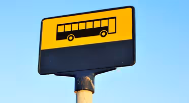 Et bussholdeplassskilt med gul bakgrunn og svart bussikon på toppen. Den nedre delen av skiltet er svart. Skiltet er montert på en stang mot en klar blå himmel.