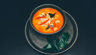 En skål med rød karrisuppe pyntet med oppskåret rød paprika og urter, plassert på en matchende mørk tallerken. Tre store grønne blader er arrangert ved siden av bollen på tallerkenen. Bakgrunnen er en jevn, mørk overflate.