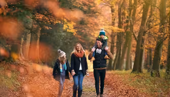 En familie på fire går gjennom en skog om høsten. Trærnes blader er oransje og brune. Faren bærer et barn på skuldrene, og moren og et annet barn går ved siden av dem. De er kledd i varme klær.