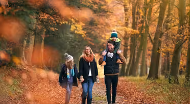 En familie på fire går gjennom en skog om høsten. Trærnes blader er oransje og brune. Faren bærer et barn på skuldrene, og moren og et annet barn går ved siden av dem. De er kledd i varme klær.