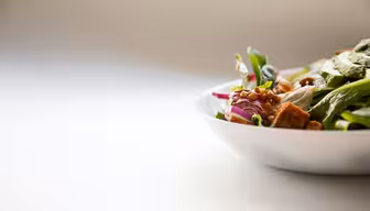 Et nærbilde av en hvit bolle fylt med en frisk salat som inneholder bladgrønt, oppskåret rødløk, linser og krutonger på en lys bakgrunn. Fokuset er på de levende ingrediensene, mens bakgrunnen er mykt uskarp.