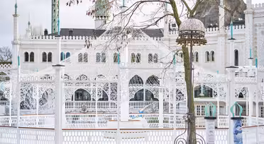 Et snølandskap med en hvit utsmykket bygning med intrikate design og kupler, muligens et palass. Bare tregrener er i forgrunnen, og scenen er innrammet av et gjerde. Himmelen er overskyet.