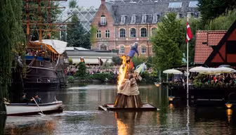 Et flytende bål med en vikingskipsfigur står i brann i en kanal, omgitt av tilskuere på båter og på en restaurant. I bakgrunnen er historiske bygninger og frodige grøntområder.