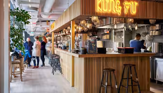 Et moderne restaurantinteriør med tredisk og barkrakker. På skiltet over står det "KUNG FU" med lysende bokstaver. Folk er tilfeldig samlet i nærheten av disken, og en person jobber på kjøkkenet. Hengende lys gir varme til innstillingen.