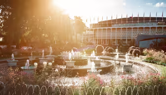 Bildet viser en solbelyst hage med en sentral fontene omgitt av blomster. En stor rund bygning med mange flagg er i bakgrunnen. Solstråler skaper en varm og innbydende atmosfære, og fremhever det livlige grøntområdet.