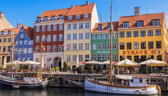 Fargerike historiske bygninger langs en kanal ved vannet i Nyhavn, København. Foran hviler to båter til kai på det stille vannet, og utekafeer med parasoller ligger langs promenaden. En klar blå himmel er over.