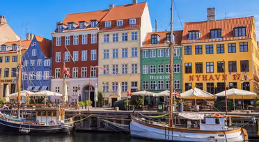 Fargerike historiske bygninger langs en kanal ved vannet i Nyhavn, København. Foran hviler to båter til kai på det stille vannet, og utekafeer med parasoller ligger langs promenaden. En klar blå himmel er over.