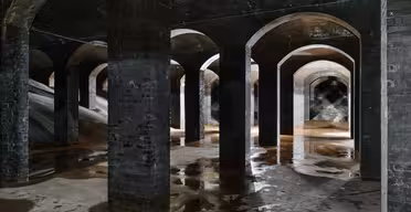 Et stort underjordisk rom med mange mursteinssøyler og buer som reflekterer svakt lys på våte gulv. Området fremstår som gammelt og industrielt, med skygger som skaper en mystisk atmosfære.