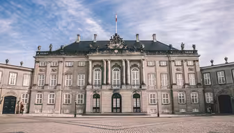 En storslått historisk bygning med en nyklassisistisk fasade, med utsmykkede statuer og store buede vinduer, står under en delvis overskyet himmel. Brosteinsgården i forgrunnen er vidstrakt og tom.