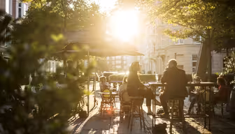 Folk sitter ved utendørs kafébord under parasoller og nyter en solrik dag. Sollyset filtrerer gjennom trærne og kaster flekkete skygger på bakken. Scenen formidler en avslappet og varm atmosfære.