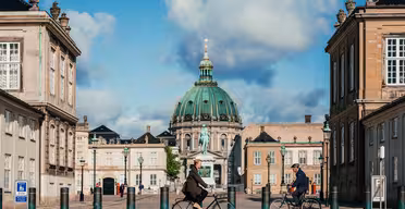 To personer på sykkel sykler mot en storslått, kuppelformet bygning innrammet av klassisk arkitektur under en blå himmel med skyer. Scenen fremkaller en følelse av bevegelse og historie i en urban setting.