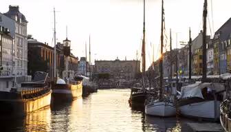 En rolig havn ved solnedgang med fortøyde båter langs begge sider av vannet. Himmelen er en varm gylden fargetone som reflekteres fra vannet. Historiske bygninger med fargerike fasader flankerer kanalen, og fullfører den pittoreske scenen.