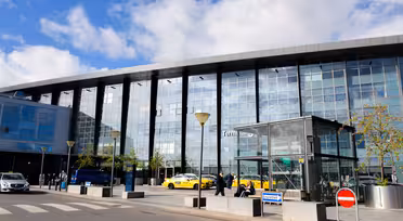 Moderne flyplassterminal eksteriør med store glassvinduer og folk som går utenfor. Noen få gule taxier står parkert ved fortauskanten. Blå himmel med spredte skyer.
