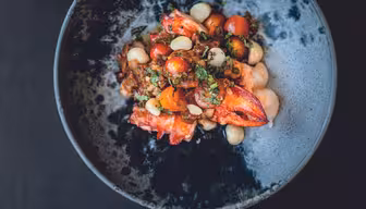 En gourmetrett servert i en mørk, teksturert bolle har en fargerik blanding av cherrytomater, hvite bønner, urter og hummerkjøtt. Ingrediensene er kunstnerisk tilrettelagt for en estetisk tiltalende presentasjon.