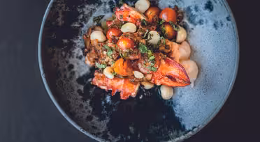 En gourmetrett servert i en mørk, teksturert bolle har en fargerik blanding av cherrytomater, hvite bønner, urter og hummerkjøtt. Ingrediensene er kunstnerisk tilrettelagt for en estetisk tiltalende presentasjon.