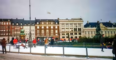 Folk skøyter på en skøytebane i urbane omgivelser, omgitt av historiske europeiske bygninger. Scenen inkluderer noen få tilskuere og nærliggende statuer, med en overskyet himmel over hodet.