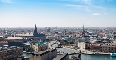 Luftfoto av et bybilde med en elv med broer, historiske bygninger og kirketårn under en klar blå himmel. Området er befolket med en blanding av moderne og tradisjonell arkitektur.