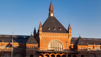 En historisk togstasjon i rød murstein med et bratt, mørkt tak og spir mot en klar blå himmel. Bygningen har store buede vinduer og et sentralt klokketårn, med sollys som lyser opp fasaden.