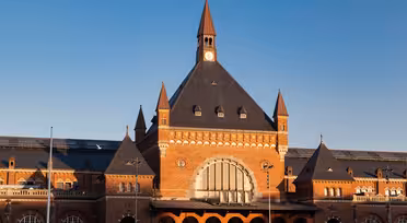 En historisk togstasjon i rød murstein med et bratt, mørkt tak og spir mot en klar blå himmel. Bygningen har store buede vinduer og et sentralt klokketårn, med sollys som lyser opp fasaden.