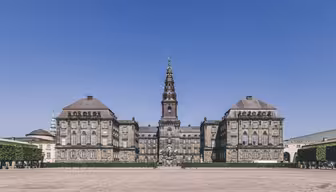 Historisk bygning med utsmykket arkitektur under en klar blå himmel, med et fremtredende tårn med et spir i midten. Rundt strukturen er en romslig brosteinsplass, foret med velstelte hekker.