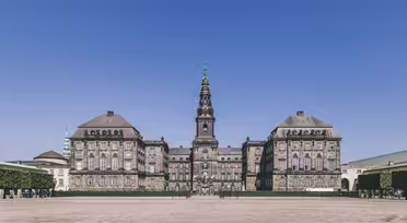 Historisk bygning med utsmykket arkitektur under en klar blå himmel, med et fremtredende tårn med et spir i midten. Rundt strukturen er en romslig brosteinsplass, foret med velstelte hekker.