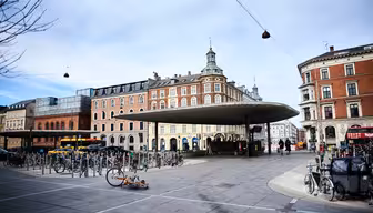 Et torg med en unik sirkulær takkonstruksjon i midten, omgitt av sykkelstativ. I bakgrunnen kan du se historiske bygninger med røde og beige mursteinsfasader. Himmelen er overskyet og noen mennesker går i nærheten.