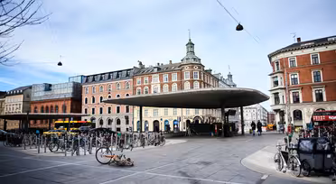 Et torg med en unik sirkulær takkonstruksjon i midten, omgitt av sykkelstativ. I bakgrunnen kan du se historiske bygninger med røde og beige mursteinsfasader. Himmelen er overskyet og noen mennesker går i nærheten.