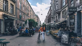 En kvinne i rød jakke går på sykkel nedover en livlig gate med utekafeer og restauranter. Folk sitter ved bordene på begge sider og nyter en solrik dag. Gaten er omgitt av murbygninger under en delvis overskyet himmel.