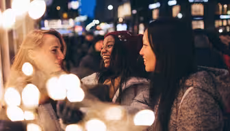 Tre kvinner prater med glede og tar en selfie i en festlig bysetting. Uskarpt lys i forgrunnen gir en varm, festlig atmosfære. Det er kveld, og den urbane bakgrunnen er mykt opplyst, noe som antyder en livlig atmosfære.