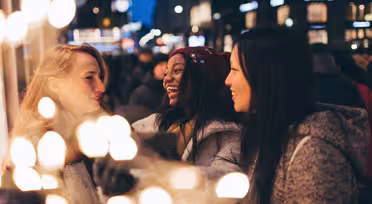 Tre kvinner prater med glede og tar en selfie i en festlig bysetting. Uskarpt lys i forgrunnen gir en varm, festlig atmosfære. Det er kveld, og den urbane bakgrunnen er mykt opplyst, noe som antyder en livlig atmosfære.