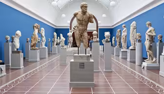 Et museumsgalleri som viser frem forskjellige eldgamle steinskulpturer på sokkel. Rommet har blå vegg og høyt buet tak med takvinduer som skaper en lys og luftig atmosfære. Besøkende ser på skulpturene.