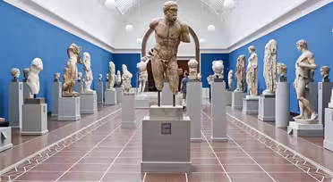 Et museumsgalleri som viser frem forskjellige eldgamle steinskulpturer på sokkel. Rommet har blå vegg og høyt buet tak med takvinduer som skaper en lys og luftig atmosfære. Besøkende ser på skulpturene.