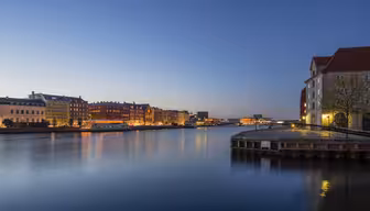 Skumringsutsikt over et rolig bybilde ved vannet med fargerike bygninger langs elven. Gatelys lyser varmt og reflekteres i det rolige vannet, mens himmelen går fra blått til dypere skumring.
