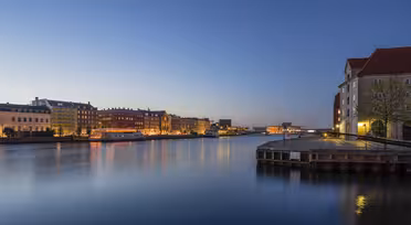 Skumringsutsikt over et rolig bybilde ved vannet med fargerike bygninger langs elven. Gatelys lyser varmt og reflekteres i det rolige vannet, mens himmelen går fra blått til dypere skumring.