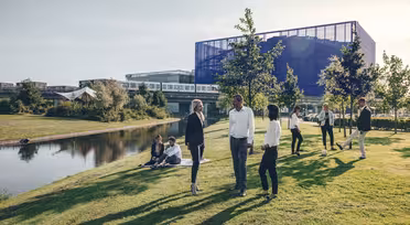 En gruppe mennesker er samlet på et gresskledd område nær en reflekterende dam. Noen står og prater, mens andre sitter ved vannet. Moderne bygninger og trær er i bakgrunnen under en klar himmel.