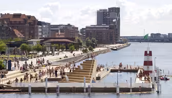 Urbant område ved sjøen med folk som nyter et stort utendørsbasseng. Bassenget er omkranset av en treterrasse med sitteplasser i lag. Mursteinsbygninger og moderne strukturer langs promenaden, med en stor vannmasse i bakgrunnen under en overskyet himmel.