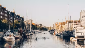 En fredelig kanalscene med flere seilbåter lagt til kai langs sidene og noen få kajakkpadlere som padler i vannet. Bakteppet har historiske bygninger under en klar blå himmel.