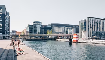 Folk soler seg på en trebrygge ved siden av en kanal på en solrik dag. Moderne bygninger er synlige i bakgrunnen. En stripet rød og hvit bøye er i vannet nær kanten av en gangbro.