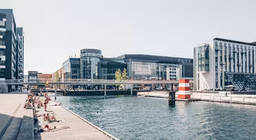 Folk soler seg på en trebrygge ved siden av en kanal på en solrik dag. Moderne bygninger er synlige i bakgrunnen. En stripet rød og hvit bøye er i vannet nær kanten av en gangbro.